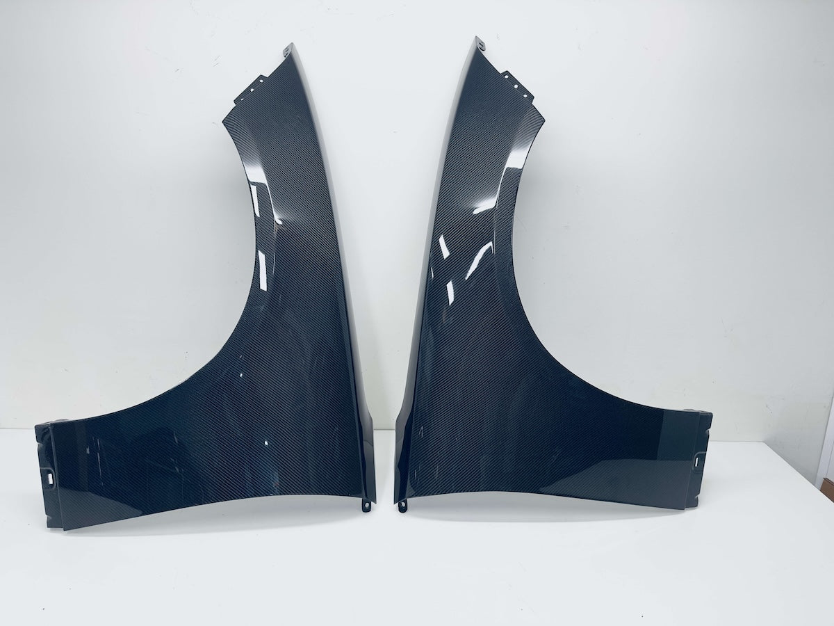 A pair of Carbon Fiber car wheel fenders displayed side-by-side on a white surface against a white wall.