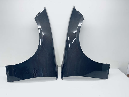 A pair of Carbon Fiber car wheel fenders displayed side-by-side on a white surface against a white wall.